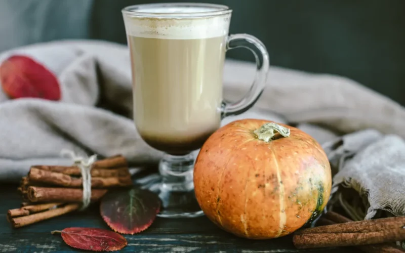 Домашно pumpkin spice лате в стъклена чаша с тиква и канелени пръчки на дървена маса