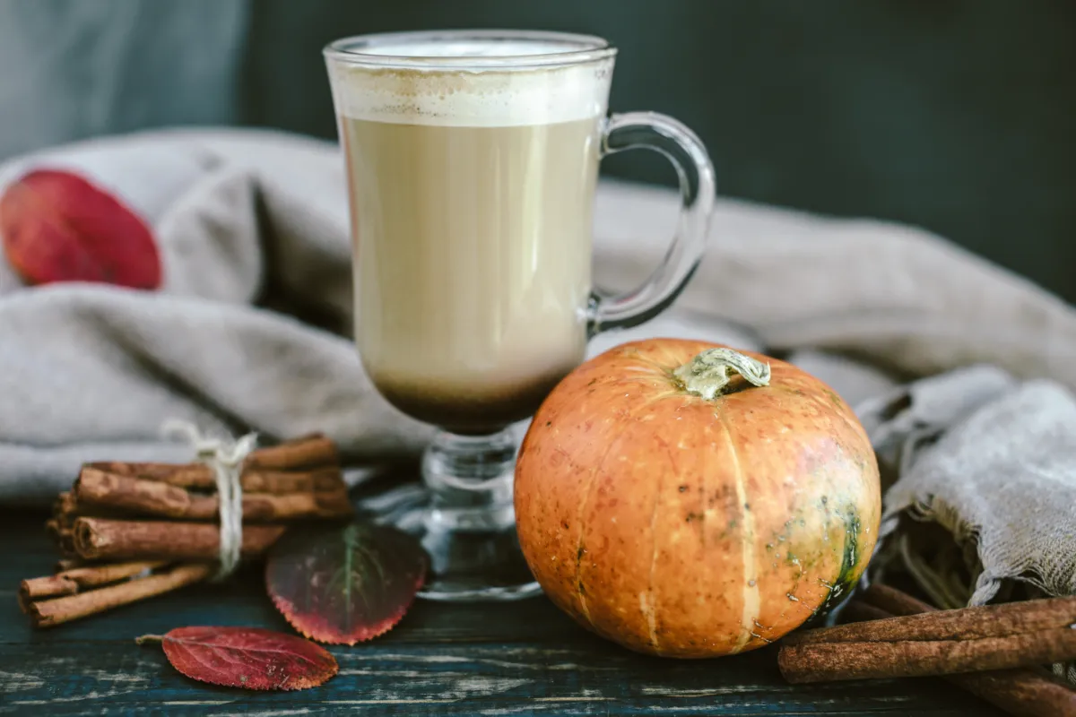 Домашно pumpkin spice лате в стъклена чаша с тиква и канелени пръчки на дървена маса